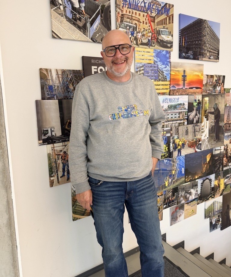 Markus Bisterfeld, neuer Bauleiter im Gerüstbau bei der Eugen Wahner GmbH, vor einer Fotowand mit Bildern verschiedener Bauprojekte.